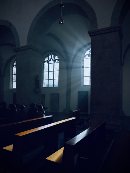 das bild zeigt das innere einer kirche. die räumlichkeit ist von hohen gewölben und steinernen säulen geprägt. durch verzierte fenster fällt tageslicht und erzeugt helle lichtstrahlen, die den dunklen raum durchdringen. im vordergrund stehen hölzerne kirchenbänke, von denen die vorderen besetzt sind. eine lampe hängt mittig von der decke. 
der obere teil des bildes hat eine kühle, bläuliche tönung, während das wenige sonnenlicht, das auf das holz der bänke fällt, warm reflektiert wird.