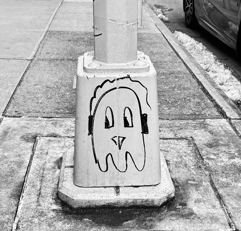 A black and white photo taken fairly close-up of the base of a large street lamp. On the base someone has drawn one of the ghosts from the Pac-Man game, wearing headphones. I don't know why it has headphones, but it's very cute. In the background is the sidewalk and a bit of a black car on the right side