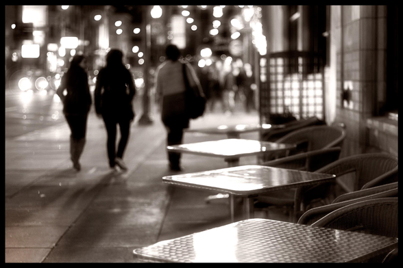 A monochrome view looking down a San Francisco street and sidewalk, at night.

On the right, a row of metal cafe tables shines... but noone is sitting there. On the left. Three people are scrolling along the sidewalk, off to enjoy an evening on this stretch of night clubs and strip joints. 

Being shot with a wide aperture, most of the scene is blurry bokeh... soft focus.