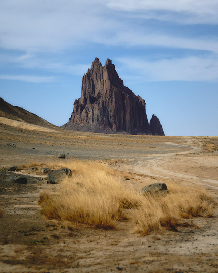 A jagged rock spire rises from a wide, arid desert plain, with tufts of dry grass and scattered stones in the foreground under a pale blue sky.