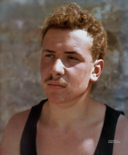 A headshot of a young light-skinned man, with curly blonde hair, and a trim mustache. He wears a black tank top, and looks into the distance, sidelit by sunlight.