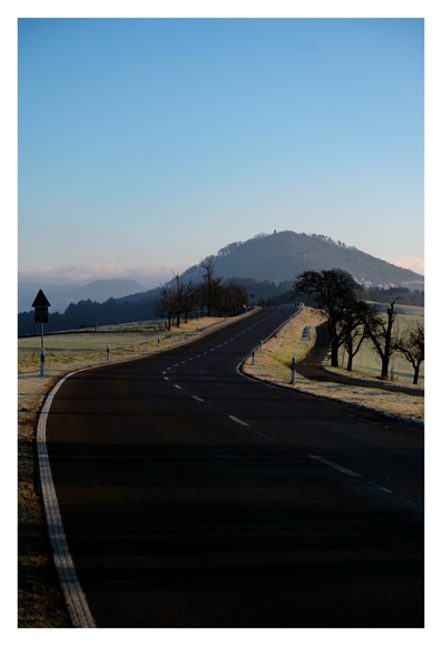 Foto im Hochformat. Blick entlang einer nach rechts geschwungenen leeren Straße. Diese führt auf einen Berg zu, der am Horizont ist. Links und rechts sind mit Frost bedeckte Wiesen und kahle Bäume. Der Himmel ist blau.