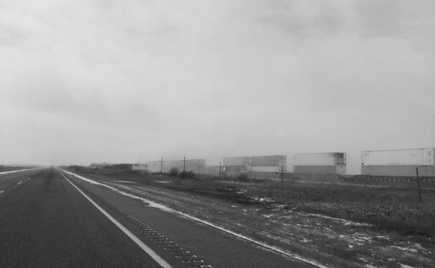 Black and white photograph taken from inside a car, of a very straight highway on the left side, the shoulder with the white line and a rumble strip, grass and a little snow in the middle, and to the right of the highway a railway with a moving train on which containers are stacked. The sky is overcast and the landscape is very flat, typical of Saskatchewan.

Photographie noir et blanc prise à bord d'une voiture, d'une autoroute très droite du côté gauche, montrant l'accottement avec la ligne blanche et une bande rugueuse, de l'herbe et un peu de neige, et à droite de l'autouroute un chemin de fer avec un train en déplacement sur lequel sont empilés des conteneurs. Le ciel est couvert et le paysage est très plat, typique de la Saskatchewan.