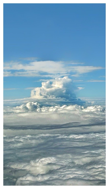 A cloudscape seen from above. White clouds completely obscure the ground far below and roll towards the horizon like a vast plain. In the distance, a towering cumulus cloud rears up like a mountain, its peaks crowned with higher, wispy clouds. Above, the sky is a clear blue.