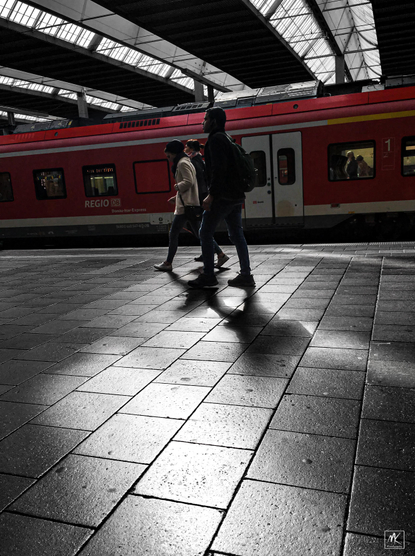 Color photo of three people walking past a red train inside a train station with sunlight coming in from roof skylights above. 