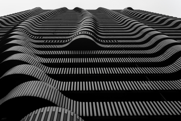 Dieses Foto zeigt die Fassade eines Hochhauses, die aus einer steilen Aufsicht fotografiert wurde. Die Architektur wirkt durch die Perspektive besonders dynamisch. Die Fassade besteht aus horizontalen, wellenförmigen Elementen, die sich rhythmisch wiederholen. Die Schwarz-Weiß-Fotografie betont die geometrischen Muster und Kontraste der Struktur. Licht und Schatten erzeugen Linien und Kurven.
 -------- 
This photograph shows the façade of a high-rise building, photographed from a steep overhead angle. The perspective makes the architecture appear particularly dynamic. The façade consists of horizontal, wave-like elements that repeat rhythmically. The black-and-white photography emphasises the geometric patterns and contrasts of the structure. Light and shadow create lines and curves.