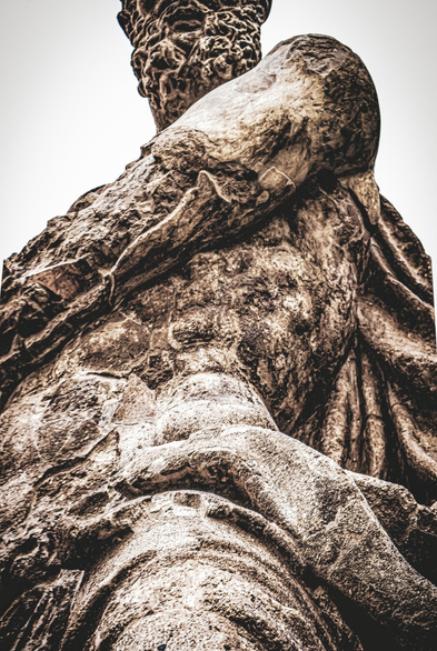 This is a low-angle, close-up view of a weathered stone statue of a man against a bright background. (Hercules)
The photograph shows the rough, textured surface of the statue, which is a mottled combination of light beige, brown, and darker grey tones. There is significant wear and tear visible in the eroded details of the stone. Hercules has a muscular physique and detailed rendering of the musculature is visible. The hair on the statue's beard is curly.