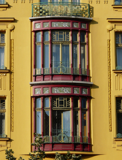 one of the centerpiece windows, it is curved outwards, dark red framing, dark naples yellow around it , gold and floral art nouveau designs around it and running through it 