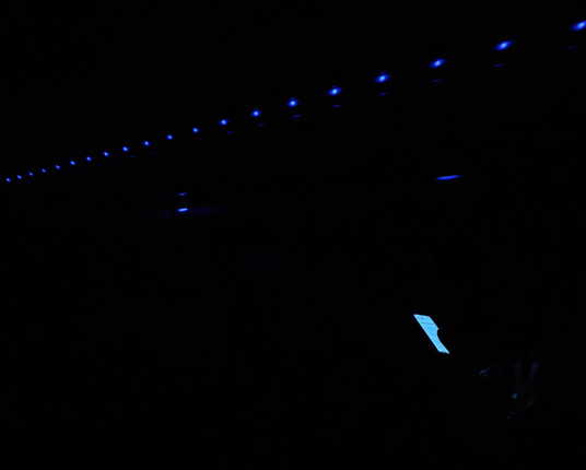 A photo taken inside a coach at night. Most of the image is black with a row of small blue lights visible overhead and a bright rectangular screen glowing blue near the lower right.