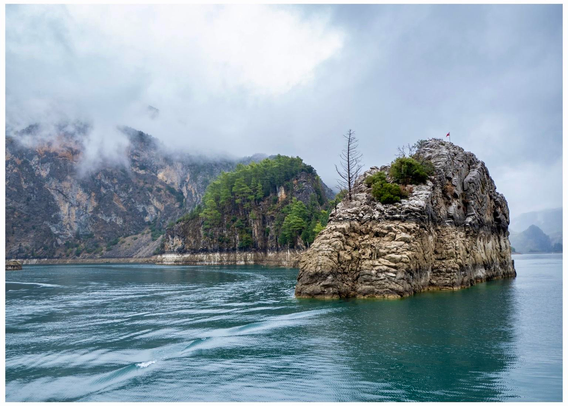 A rocky island rises from calm, turquoise waters, surrounded by lush green trees and steep, mist-covered mountains. The sky is cloudy, adding a serene atmosphere to the landscape. Taken in the Green Canyon, Turkey