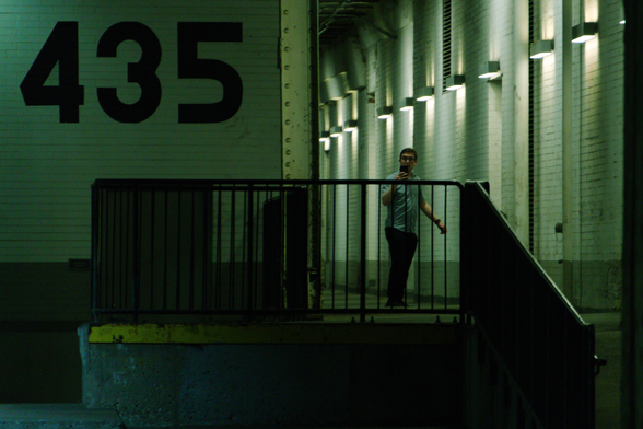 A man rounds a corner in a loading dock, staring out into the open, camera in outstretched hand, looking like he might lost or anxious. A green light bathes the scene in discomfort. The numbers 435 are painted on the brick wall to the man's left, about the same size as he is.