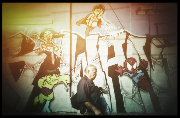 A man, with eyes closed and head slightly down-turned, walks past a mural of cartoon DC and Marvel action figures. He is burdened down with packages, totally lost in thoughts or daydreams while Hulk, Wonder Woman, Superman, and Spiderman fight around him.