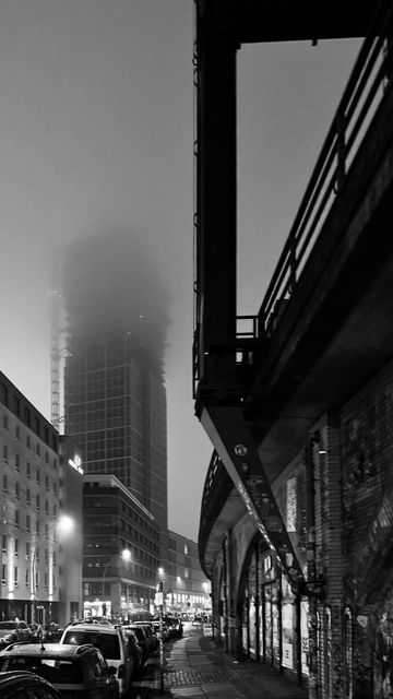 A foggy urban scene in black and white, showcasing a partially obscured tall building in the background. In the foreground, a street lined with parked cars is visible, alongside a brick wall with graffiti. An elevated structure looms overhead, adding to