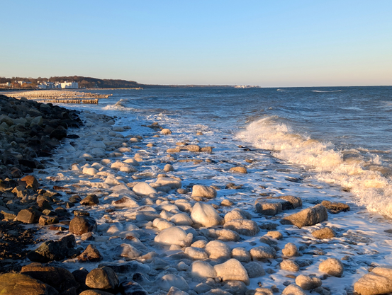 Der Betrachter steht parallel zu einem mit großen Steinen bedeckten Strand. Links erhebt sich ein Damm aus Steinen. In der Bildmitte sind die im flachen Wasser liegenden Steine mit Eis überzogen. Rechts rollt die Brandung der Ostsee heran. Im Hintergrund vereiste Buhnenreihen und die Häuser von Heiligendamm. Am Horizont ist Kühlungsborn zu erkennen. Blauer Himmel.