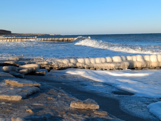 Im Vordergrund ein vereister Strand, an dem kleine Eisschollen liegen. Eine dick mit Eis überzogene Buhnenreihe führt nach rechts in die Brandung. Weiter hinten mehrere weitere Buhnenreihen und eine Seebrücke. Am Horizont Häuser.
