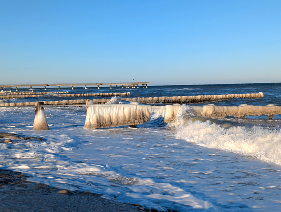 In der schäumenden Brandung stehen mehrere Holzpfähle, die mit einem Edelstahl-Handlauf verbunden sind. Von diesem hängen kleine Eiszapfen und zum Teil dicke vorhangartige Eisgebilde heran. Weiter hinten vereiste Buhnenreihen und eine Seebrücke.