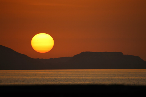 Sunset over the sea in Heraklion, Greece. The large, orange sun is low on the horizon, painting the sky in warm hues. In the foreground, the sea appears calm, and in the background, dark hills are silhouetted.