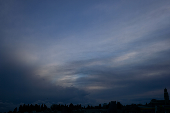 Photo looking up at a cloud-streaked sky, it gets dark towards the lower-left and lower-right corners. Across the very bottom are silhouettes of low-rise buildings, including at least one dome and one domed tower.