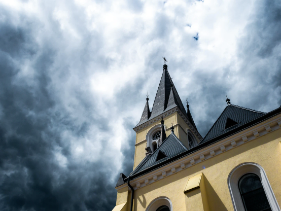 Kirchturm, Teil des Daches und der Wand einer gelb gestrichenen Kirche von unten hoch fotografiert. 
Der Himmel liefert Drama.  