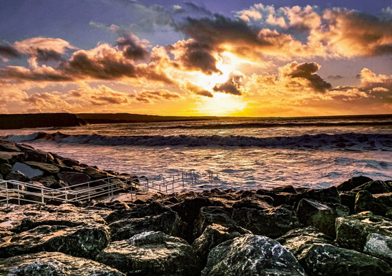 The foreground features a collection of large, dark rocks, some of which appear wet. There's a white metal railing and steps descend from the rocks towards the ocean. The ocean stretches out towards the horizon with visible waves, some crashing and creating white foam.
The sky is filled with a mix of dark blue and vibrant orangey/yellow clouds, illuminated by the setting sun behind them. The sun peeks through a gap in the clouds, casting a golden glow across the water and sky. In the distance, a dark landmass is visible on the horizon.