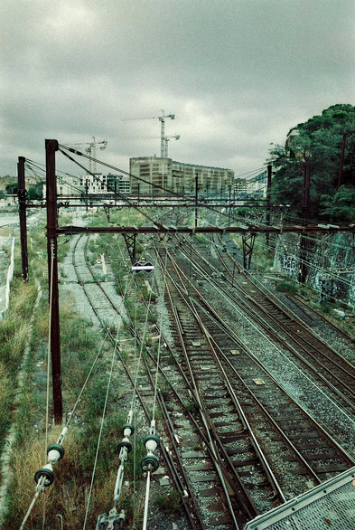 Enchevêtrement urbain, des rails de train au premier plan se dirigeant vers des bâtiments en construction avec grues au second plan.