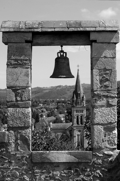Black and white photograph of a church steeple in a rural landscape of hills, trees and houses, taken from above through a stone frame in which a metal bell is fixed.

Photographie noir et blanc du clocher d'une église dans un paysage rural de collines, d'arbres et de maisons, prise en surplomb à travers un cadre en pierres dans lequel est fixée une cloche en métal.