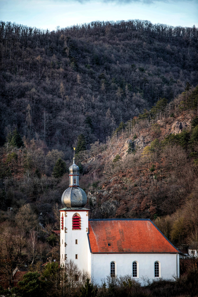Das Bild zeigt eine kleine Kirche im barocken Stil vor einem steilen, bewaldeten Berghang. Das Kirchengebäude hat weiß verputzte Wände, ein Satteldach mit roten Ziegeln und einen Glockenturm, der mit einer silberfarbenen Zwiebelkuppel und einem goldenen Kreuz abschließt. Der Turm verfügt über kleine, rot umrandete Fenster. Die Hauptfassade des Kirchenschiffs hat ebenfalls mehrere schmale, hohe Fenster mit Rundbögen.
Der umgebende Hang ist dicht bewaldet, wobei die meisten Bäume kahl sind und in dunklen Braun- und Grautönen erscheinen, was auf Spätherbst oder Winter hindeutet. Der Boden ist felsig und mit trockenem Laub bedeckt. Die Komposition hebt den Kontrast zwischen dem hellen, architektonisch klaren Bauwerk und dem dunklen, wilden Naturhintergrund hervor.