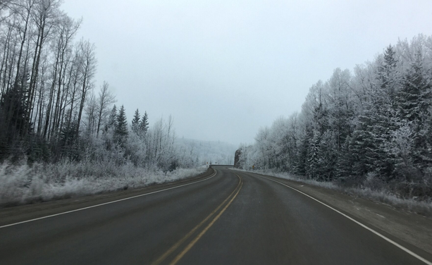 Photograph of an asphalt road in a winter landscape, taken from a car. The road is clear, with a yellow line down the center and white lines on either side. It curves to the right in the distance, in the center of the image, to skirt a rock face. On either side, the forest is completely frosted. The sky is hazy and opaque. The frost and mist give the landscape a bluish-white tint.

Photographie d'une route asphaltée dans un paysage hivernal, prise à bord d'une voiture. La route est bien dégagée, avec une ligne jaune au centre et des lignes blanches de chaque côté. Elle se courbe vers la droite au loin, au centre de l'image, pour contourner un mur de roche. De chaque côté, la forêt est complètement givrée. Le ciel est brumeux et opaque. La givre et la brume donnent une teinte blanc-bleutée au paysage.