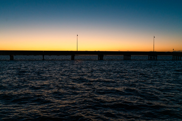 The sun sets over the Charlotte Harbor near the Gulf of Mexico in Florida. The bridge is silhouetted by the radiant orange horizon. The waters are a deep blue.