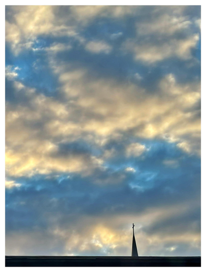 A sky full of clouds just after sunrise, brushed with both shadows and golden light. At the bottom of the frame is the rooftop edge of a nearby building, and beyond that, the silhouette of a distant church steeple with a cross, dark against the golden clouds.