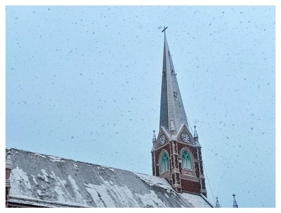 The roof and bell tower of a small church on a snowy day. The angled roof extends from left to just past center frame, where the steeple rises and tapers to a cross at its apex. On the two visible sides of the tower are teal windows with horizontal slats, set into arched frames; above those are circular clocks with Roman numerals, the hands indicating 9:00. The upper surfaces of the building are all dusted with snow, as more snowflakes fill the gray sky.
