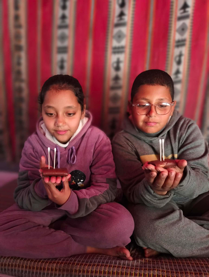 The twins, Aylan in a purple hoodie and sweatpants and Elias in a gray hoodie and sweatpants, sit cross-legged holding small, rectangular treats with cotton swabs inserted, looking down at them. They are in front of a red and white striped background.