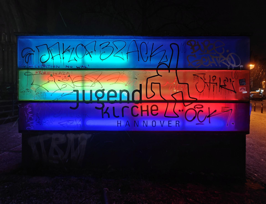 A photo of an illuminated outdoor sign reading “Jugendkirche Hannover” (Youth Church Hannover) with horizontal bands of blue, green, and red light. The sign is covered with scribbles and graffiti tags.