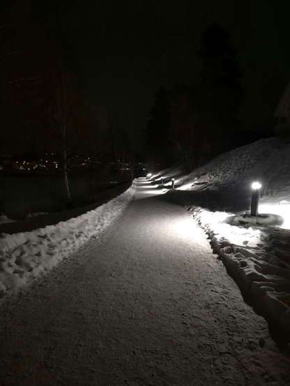 A promenade along a river in the dark. There is snow on the ground, but the path has been cleared and the snow has piled up on both sides of the path. Small lamps on one side of the path illuminate the way, and in their glow you can still see some of the trees standing along the path.