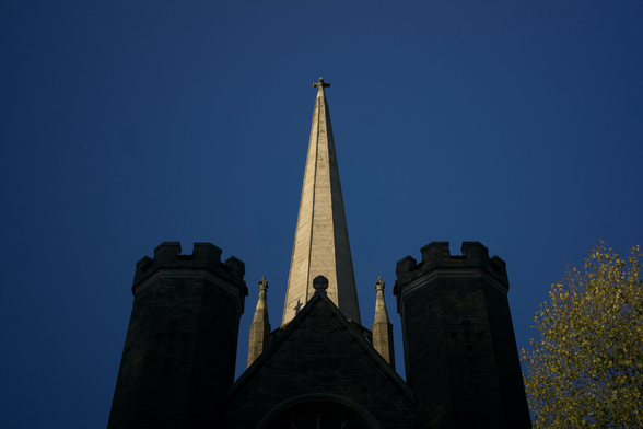 Photo of an extremely pointed spire in bright daylight, against a clear deep blue sky, behind near-silhouettes of a couple of crenellated octagonal towers either side of the end of a pitched roof, which sort of look like engines on a space rocket, or an X- or Y-wing from Star Wars, if you're that way inclined.