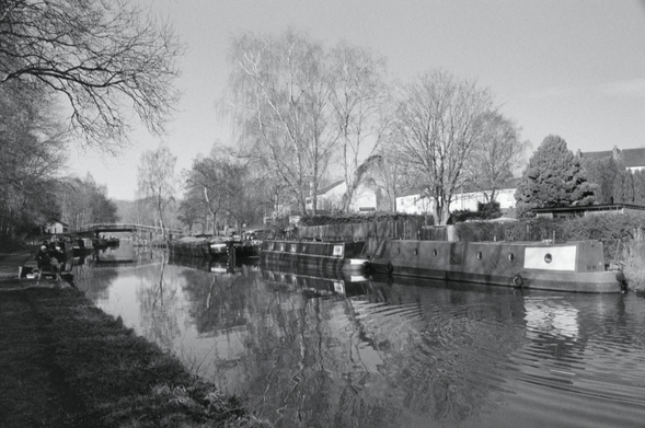 A black and white 35mm film photo of a fisherman on the towpath, narrowboats and reflections on the Leeds & Liverpool Canal at Crooke near Wigan.