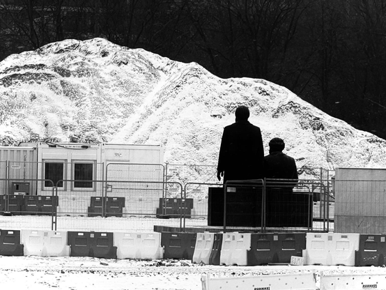 A black and white image depicting two statues, one seated and one standing, in front of a snowy mound. Surrounding them are construction barriers and equipment, creating a stark and somber atmosphere.