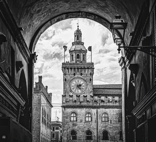 This is a black and white image showing a clock tower (Il torre dell'Orologio, part of Palazzo d'Accursio) through an archway. The archway it located in Via delle Pescherie Vecchie. It Is a medievally constructed building made out of stone. There are windows visible. At the very top there are two people on the balcony looking out.