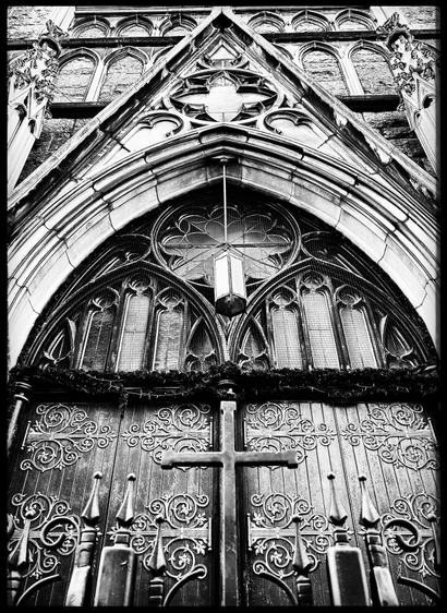 A black-and-white photo in fairly extreme close-up. The center of the photo shows a large iron cross that is part of the gate that closes in front of the church steps. Two points from the gate can be seen on either side of the cross and the doors behind the cross have ornate curlycue iron work on the large wooden panels. Above that is a little greenery and two pointed arches over each door with three panels inside each one. Above them in the middle is a large circle with stained glass work. All of that is set within a large white stone pointed arch way as well. That comes to a point on top with some more decorative work on the face. There is a light hanging down in between the two door panel archways that is directly over the top of the cross. It was a difficult photo because the beautiful old church is surrounded by very tall glass and steel buildings as well as an on-ramp with lots of traffic whizzing by