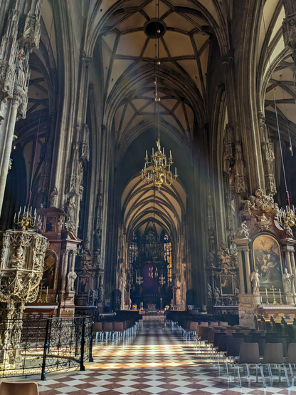 The majestic Gothic interior of St. Stephen's Cathedral in Vienna, Austria, featuring soaring ribbed vaults, ornate columns with statues, and a distinctive red and white checkerboard marble floor. The image shows rows of modern chairs leading towards the high altar, illuminated by light streaming through windows.