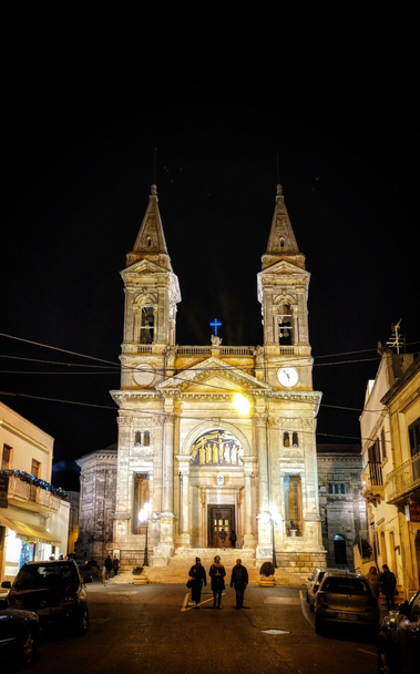 This image captures a beautifully illuminated church facade at night. The church features a grand, symmetrical design with two prominent bell towers flanking a central entrance. Each tower is adorned with a clock face and topped with a pointed spire, adding to the architectural elegance of the structure. The facade is intricately detailed, with columns, arches, and decorative elements that highlight its historical and artistic significance.

The church is bathed in warm, golden lighting, creating a striking contrast against the dark night sky. The surrounding area appears to be a quiet town square, with a few people walking in front of the church and parked cars lining the street. The adjacent buildings, also softly lit, contribute to the charming and serene atmosphere of the scene.