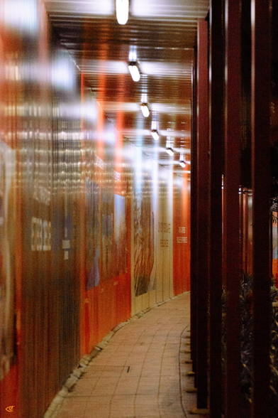 Narrow curved corridor with a tiled floor, corrugated metal construction walls on the left (partly blurred), dark support posts on the right, and a row of fluorescent ceiling lights receding into the distance.