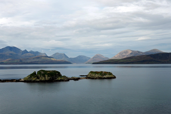 Blick auf eine Bergkette, die Cuillin Hills in Schottland auf der Isle of Skye. Im Vordergrund kleinere Inseln im Loch Eishort. Der Himmel ist grau, wolkenverhangen. 

View of a mountain range, the Cuillin Hills in Scotland on the Isle of Skye. In the foreground are smaller islands at Loch Eishort The sky is grey and overcast. 