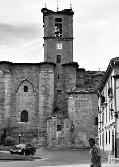 La imagen en blanco y negro muestra una iglesia de piedra con una torre campanario que tiene un reloj en su fachada. En la parte superior de la torre se observan campanas y nidos de cigüeña. A la derecha hay casas adosadas con balcones y faroles, y en la esquina inferior un hombre camina por la calle mientras un coche está estacionado cerca de la acera.