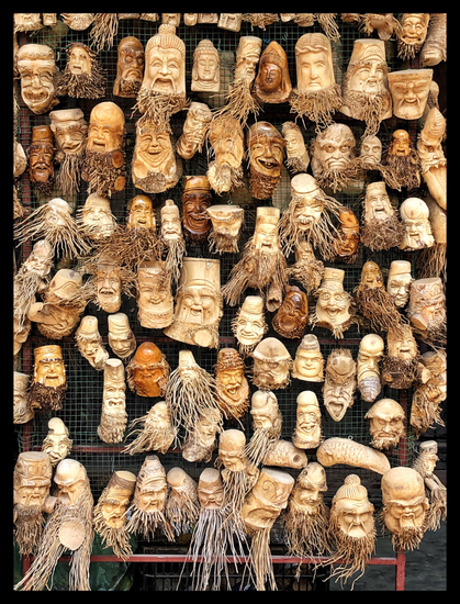 A crowded display of miniature wooden sculptures hung on a mesh screen. The sculptures are made from tree roots and mostly depict the faces of old men with exaggerated smiling or grimacing expressions, many sporting tangled “beards” of root tendrils.