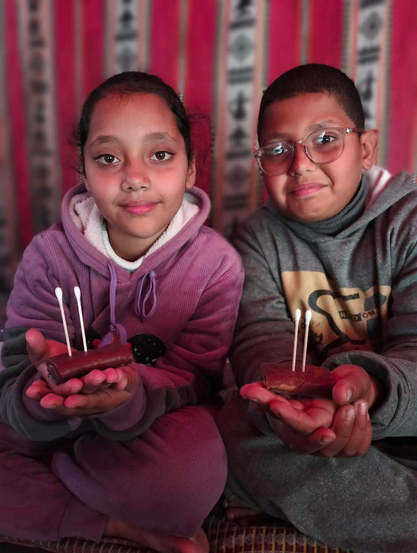 The twins, Aylan & Elias, smiling at the camera, each holding a chocolate bar with two cotton swabs inserted, set against a red-and-white striped background. The girl, wearing a purple hoodie, has dark hair and brown eyes. The boy, wearing glasses and a gray hoodie, looks at the camera.