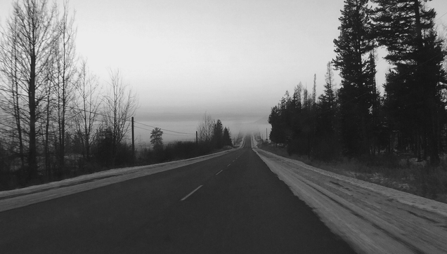 Black and white photograph taken from a car, of an asphalt road that descends steeply towards a mist-covered lake. Trees line the road on both sides, as well as poles and power lines, and the shoulders are covered in ice.

Photographie noir et blanc prise depuis une voiture d'une route asphaltée qui descend fortement vers un lac couvert de brume. Des arbres bordent la route de chaque côté, ainsi que des poteaux et des lignes électriques, et les accottements sont couverts de glace.