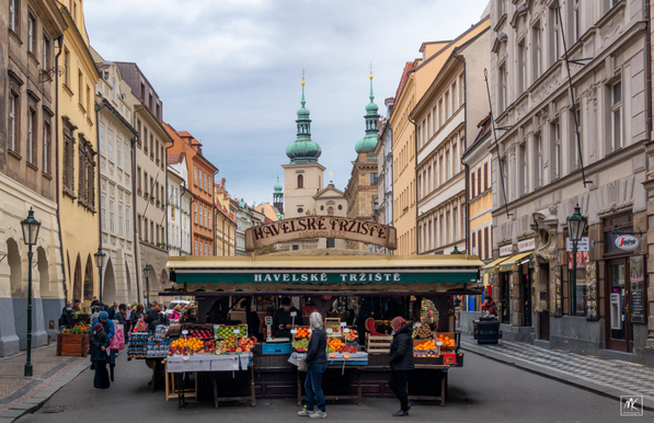 Color photo of the end of a long low market stall structure located in the center of a street in Prague. There are a couple of shoppers stopped to look at the displayed fruit for sale. Down the street in the background are twin greenish onion dome and spire topped towers of a church.
