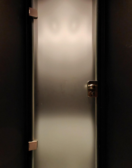 A photo of a frosted glass door, with shiny metal hinges, a door handle and lock. Dark walls frame the door with bright light behind the glass.