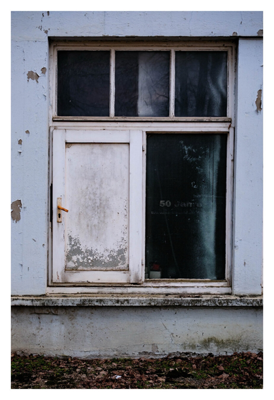 Foto im Hochformat. Ein altes heruntergekommenes Fenster in einer Hauswand mit aufgeplatztem hellblauem Putz. Das Fenster ist nur ca. 50 cm über dem Boden und besteht aus zwei rechteckigen Öffnungen, darüber drei kleinere Rechtecke. Das linke untere Fenster ist eine weiße Tür mit aufgeplatztem Holz. Die Türe ist insgesamt nur ca. einen Meter hoch, also sehr klein.